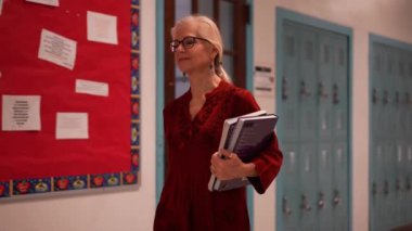 Okulda boş bir koridorda kameraya doğru yürüyen mutlu ve olgun bir kadın öğretmenin yavaş çekimde portresi..