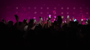 Seyirci kitlesinin yavaş çekim geniş açılı görüntüsü. Elleri havada, telefonları havada, siluetlerini sallayarak bando sahnesinde, parlak mavi, flaş ışıkta ilerliyorlar. Festivaldeki mutlu insanlar müziğin tadını çıkarıyor