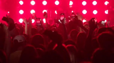 Seyirci kitlesinin yavaş çekim geniş açılı görüntüsü. Elleri havada, telefonları havada, siluetlerini sallayarak bando sahnesinde, parlak mavi, flaş ışıkta ilerliyorlar. Festivaldeki mutlu insanlar müziğin tadını çıkarıyor