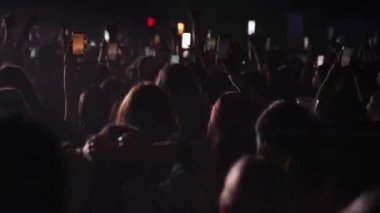 Seyirci kitlesinin yavaş çekim geniş açılı görüntüsü. Elleri havada, telefonları havada, siluetlerini sallayarak bando sahnesinde, parlak mavi, flaş ışıkta ilerliyorlar. Festivaldeki mutlu insanlar müziğin tadını çıkarıyor