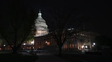 Ağaçların arasından geçen kamera, gece Washington DC 'de mavi mor gökyüzüne karşı sallanan ışık ve bayraklarla ABD Kongre Binası' nı gözler önüne seriyor..