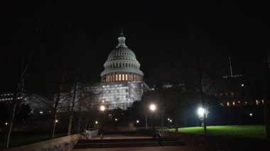 Kamera, gece mavi mor gökyüzüne karşı Washington DC 'de dalgalanan ışık ve bayraklarla, Amerikan Kongre Binası' nın batı tarafında inşaat iskelesini gösteriyor..