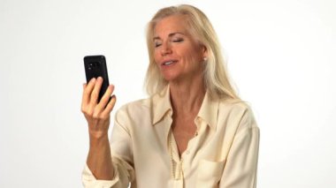 50 'li, 60' lı yaşlarda, mutlu ve olgun bir kadın. Sarışın, beyaz arka planda, modern iç mekanda izole edilmiş akıllı telefondan görüntülü konuşma yapıyor. Arkadaşlarla, ailelerle, iş arkadaşlarıyla konuşma kavramı.