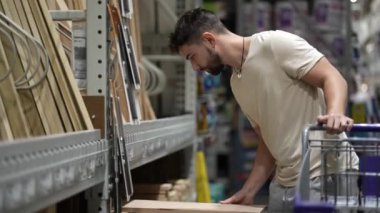 Sakallı genç bir adam hırdavatçıda ya da depoda kereste seçiyor..