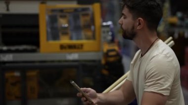 Hırdavatçı dükkanının kereste reyonundaki telefona bakarken alışveriş arabasını iten adamın yan görüntüsü.