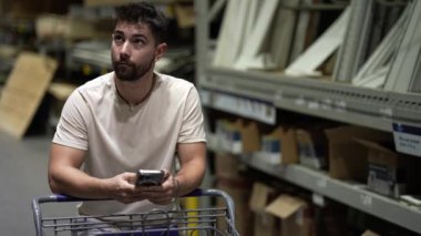 Genç adam ahşap bölmedeki hırdavatçı dükkanında telefon kullanarak alışveriş arabasına yaslanıyor..