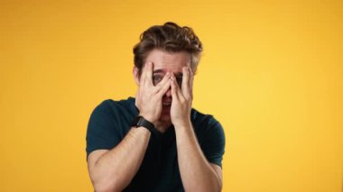 Kameraya bakarken şaşırmış bir yüz ve eller saklanıyor. 20 'li yaşlardaki genç adam sarı arka plan stüdyosunda izole edilmiş. Portre samimi duygular yaşam tarzı konsepti.