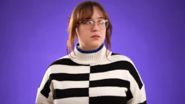 Kameraya bak ve sessiz ol de. Parmaklarını dudaklarına koy. Gözlüklü ve kazaklı genç bir kadın stüdyoda mor arka planda izole edilmiş. Samimi duyguların portresi, yaşam tarzı konsepti.