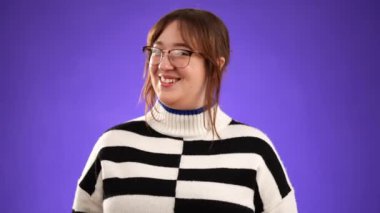 Gülümseyen güzel bir kızın portresi 20 'li yaşlarda mutlu bir kadının gözlük takması ve kazak giymesi, bana sorması çok tatlı. Ellerini göğsüne koy, mor arka plan stüdyo resmine izole edilmiş..