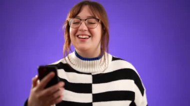 20 'li yaşlarda, süveter giyen bir kadın telefonda bir arkadaş veya doktorla görüntülü konuşma yapıyor. Mor arka planda izole edilmiş bir web kamerası var..