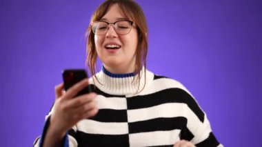 20 'li yaşlarda, kazak sallayan ve telefonda bir arkadaş veya doktorla görüntülü konuşma yapan bir kadın, mor arka planda izole edilmiş bir web kamerası.