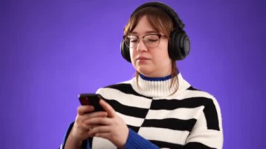 Telefonda müzik dinleyen, kulaklık takan, mor arka plan stüdyosunda yalnız poz veren mutlu genç kadın portresi..