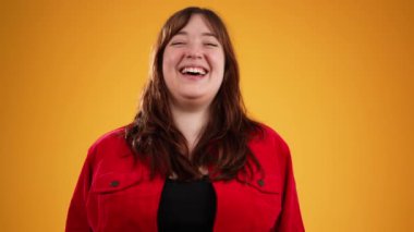 Kahverengi saçlı, kırmızı ceketli, gülen ve işaret eden genç bir kadın. Sarı arka planda izole edilmiş, fotokopi alanı ağır çekimde.