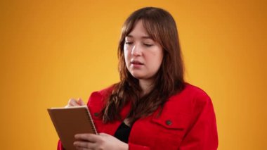 Düşünceli gazeteci kadın notlar alıyor, not defteri günlüğüne kalemle düşünceler yazıyor, liste yapmak için, iyi fikir. Güzel esmer kız, sarı stüdyo arka planında izole edilmiş.