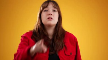 Buraya gel, beni takip et! Kırmızı ceketli güzel uzun saçlı bir kız tek parmakla işaret ediyor, yaklaşmaya davet ediyor, şakacı bir flört gibi görünüyor. Sarı arkaplanda izole edilmiş