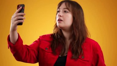 20 'li yaşlarda mutlu, kırmızı ceketli bir kadının portresi cep telefonuyla selfie çekiyor sarı arka plandaki stüdyoda hoş sohbetler yapıyor..