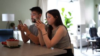 Kamera, mutfak tezgahında oturan genç bir çifti görüntülemek için geri çekiliyor. Telefonla mesajlaşıyorlar, evde çalışıyorlar..