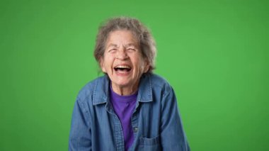Gülümseyen mutlu, yaşlı bir kadının yavaş çekimde portresi. Kırışık cilt ve yeşil perde arkasında izole edilmiş gri saçlar..
