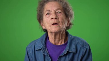 Gülümseyen dişsiz, yaşlı, buruşuk derili ve gri saçlı yaşlı kadının komik, yavaş çekim portresi yeşil ekran arka plan stüdyosundan izole edilmiş bir sırrı dinlemek için kulaklarını tıkıyor.