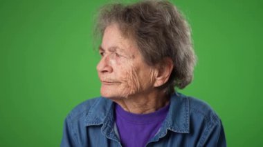 Sinirli, sinirli, dişsiz, yaşlı yaşlı bir kadının yavaş çekimde çekilmiş portresi. Sessiz, kırışık derili, yeşil ekran arka planında gri saçları olan yaşlı bir kadın..