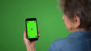Yeşil ekran arkaplanında izole edilmiş yeşil ekran izleme noktaları olan akıllı telefonla konuşan yaşlı bir kadının omuz manzarası üzerinde. Boş ekran ile cep telefonunu tut