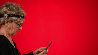 Akıllı telefonuyla kayarken komik yaşlı kadın karede yürür durur, durur, gülümser ve karede yürümeye devam eder. Kırmızı arkaplanda izole