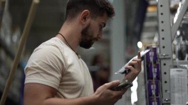 Sakallı bir adamın nalbur deposundaki eşyaları telefon kullanarak karşılaştırırken yakın görüntüsü..