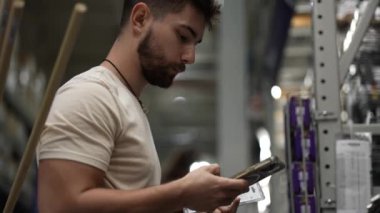 Sakallı bir adamın nalbur deposundaki eşyaları telefon kullanarak karşılaştırırken yakın görüntüsü..