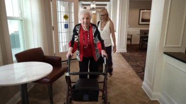 Gülümseyen olgun bir kadın huzurevinde ya da huzurevinde Walker kullanarak yaşlı bir kadınla yürüyor..