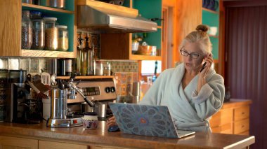 Telefonla konuşan ve dizüstü bilgisayarla çalışan bir kadın mutfak ortamında bornoz giyip gülümsüyor..