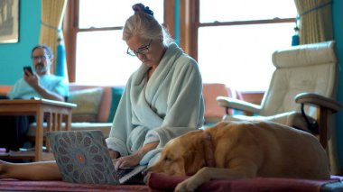 Bornozlu olgun bir kadın dizüstü bilgisayarda yanında köpek ve telefondaki adamla çalışıyor..