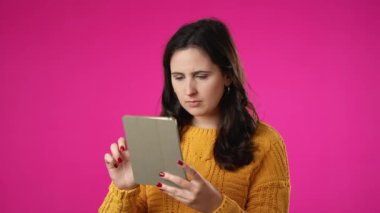 Mutlu gülümseyen genç kadın portresi 20 'li yaşlarda kazak alışverişinde tablet bilgisayarda parmakla işaret ederek pembe arka plan stüdyosunda izole edilmiş..