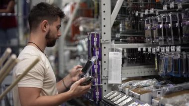 Sakallı bir adam nalbur deposunda eşya aramak için telefon kullanıyor..