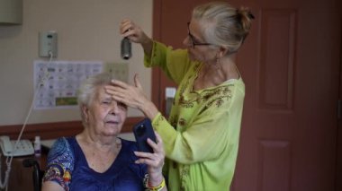 Yüzü morarmış yaşlı bir kadının yakın çekimi. Rehabilitasyon merkezinde aile üyesi tarafından saçı taranıyor..