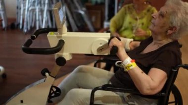 Kolu kırık yaşlı bir kadın, rehabilitasyon merkezinde bisiklet sürüyor. Sarı kol bandında düşme riski.