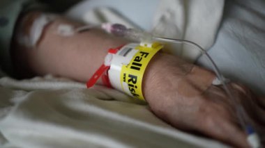 Hastane yatağında, sarı bir sonbahar bileziği takmış ve serum takmış hastaların yakın plan fotoğrafları. Hastanedeki hastaların kırılganlıklarını ve savunmasızlıklarını vurguluyorlar..