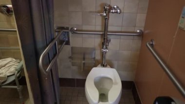 Beyaz tuvaletli, parmaklıklı ve duş perdeli bir banyo. Klozetin kapağı yükseltilmiş ve gidonu da kolay erişim için yerleştirilmiş.. 