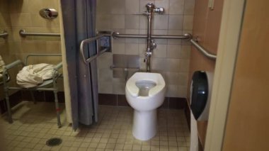 Erişilebilirlik için tasarlanmış bir banyoda musluğu olan bir lavabo, tutacağı olan bir tuvalet ve duş başlığı ve kontrol düğmesi olan bir duş bulunur. Banyoda fayanslı duvarlar ve duş perdesi var..