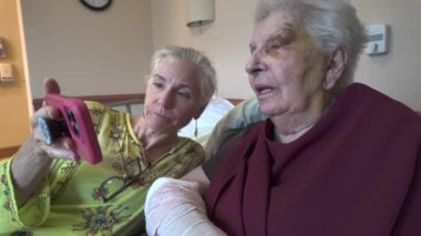 Sarı saçlı genç bir kadın kolunda bandaj ve gözünün etrafında çürük olan yaşlı bir kadına pembe bir akıllı telefon gösteriyor..