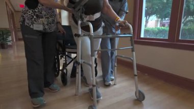 Tekerlekli sandalyedeki bir kişiye ve yürüteç kullanan bir başkasına hastane koridorunda bir personel tarafından yardım ediliyor.. 
