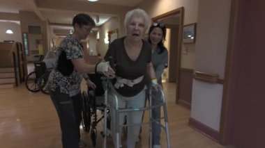 Olney, MD, ABD - 10 13 2024: Elinde bandaj olan yaşlı bir kadına huzurevi koridorunda yürüyen bir kadınla yürümesi için iki kadın yardım ediyor. Arka planda tekerlekli sandalye var..