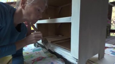 Sarı saçlı bir kadın ahşap bir dolabı boya fırçasıyla beyaza boyuyor. O, mavi bir gömlek giyiyor ve yerde diz çöküyor. Dolabın bir rafı ve altında küçük bir açıklığı var..