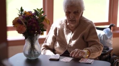 Beyaz saçlı yaşlı bir kadın tekerlekli sandalyede oturuyor ve masada kağıt oynuyor. Önplanda çiçekli bir vazo var..