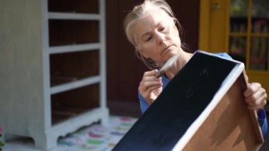 Gri saçlı bir kadın siyah ahşap bir çekmeceyi beyaz boyayla boyuyor. O, sağ elinde bir fırça tutuyor ve göreve odaklanıyor. Arka planda beyaz bir dolap var..