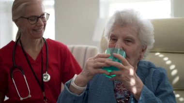 Kırmızı önlüklü bir bakıcı, susuzluğun önemini vurgulayarak, iyi ışıklandırılmış kapalı bir mekanda yaşlı bir kadının deniz mavisi bardaktan su içmesine yardım ederken gülümsüyor..