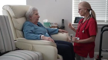 Bakıcı, bir sağlık merkezindeki rahat bir sandalyede oturan yaşlı bir kadına özen gösterir ve bağlantıyı ve konforu teşvik eder..