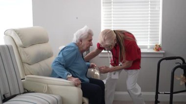 Bir bakıcı, yaşlı bir kadının önünde oturur. Parlak bir sağlık tesisinde bağlanma ve bakımı teşvik eder ve etkileşim için sıcak bir ortam yaratır..