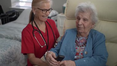 Bir sağlık görevlisi, rahat bir odada otururken, sakin bir ortamda kişisel bakım ve ilgi gösteren, uzaktan kumandalı yaşlı bir kadına yardım eder..