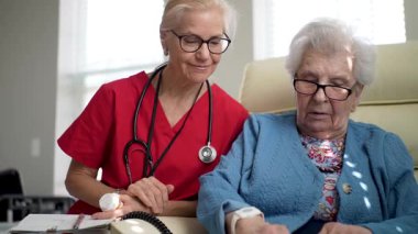 Kırmızı önlüklü bir bakıcı açık mavi hırkalı yaşlı bir kadınla sıcacık bir şekilde yakınlaşıyor ve sağlık hizmetleri ortamında bilişsel etkileşimi teşvik ediyor..