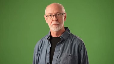 Gri saçlı ve gözlüklü yaşlı bir adam, portresindeki duyguların bir karışımını yansıtan, katı yeşil bir arka planda sakince duruyor. Anlayış gösteriyorum, kafa sallıyorum..